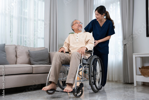 A young Asian female nurse or physical therapist in a blue uniform, standing and talking happily with a senior Asian man sitting on a wheelchair while pushing him to relax in the living room at home.