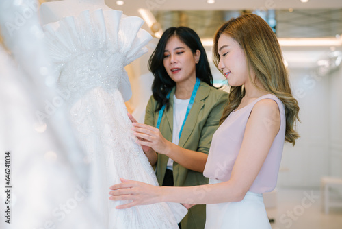 Asian bride is smiling while choosing wedding dress in modern wedding salon, Attractive designer girl using tape meter fitting on wedding dress at wedding studio