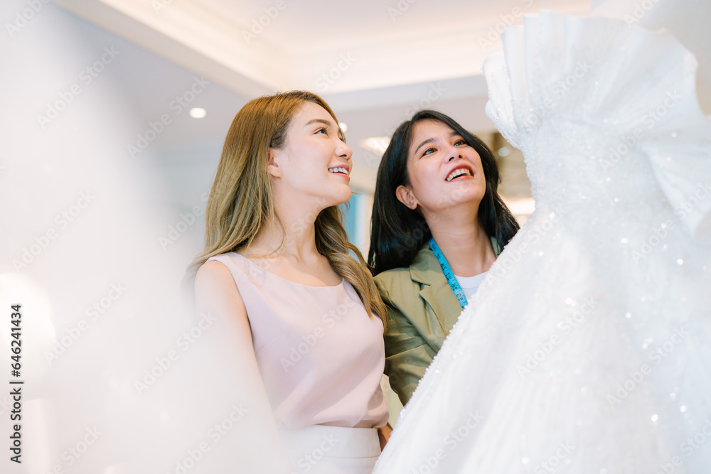 Asian bride is smiling while choosing wedding dress in modern wedding salon, Attractive designer girl using tape meter fitting on wedding dress at wedding studio Asian bride is smiling while choosing wedding dress in modern wedding salon, Attractive designer girl using tape meter fitting on wedding dress at wedding studio
