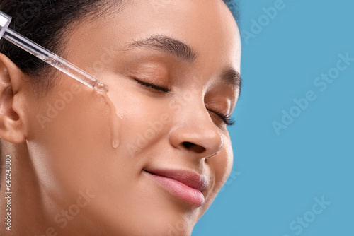 Beautiful woman applying serum onto her face on blue background, closeup. Space for text