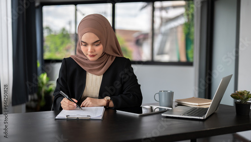 A focused Asian Muslim businesswoman is filling out forms or signing documents at her desk.