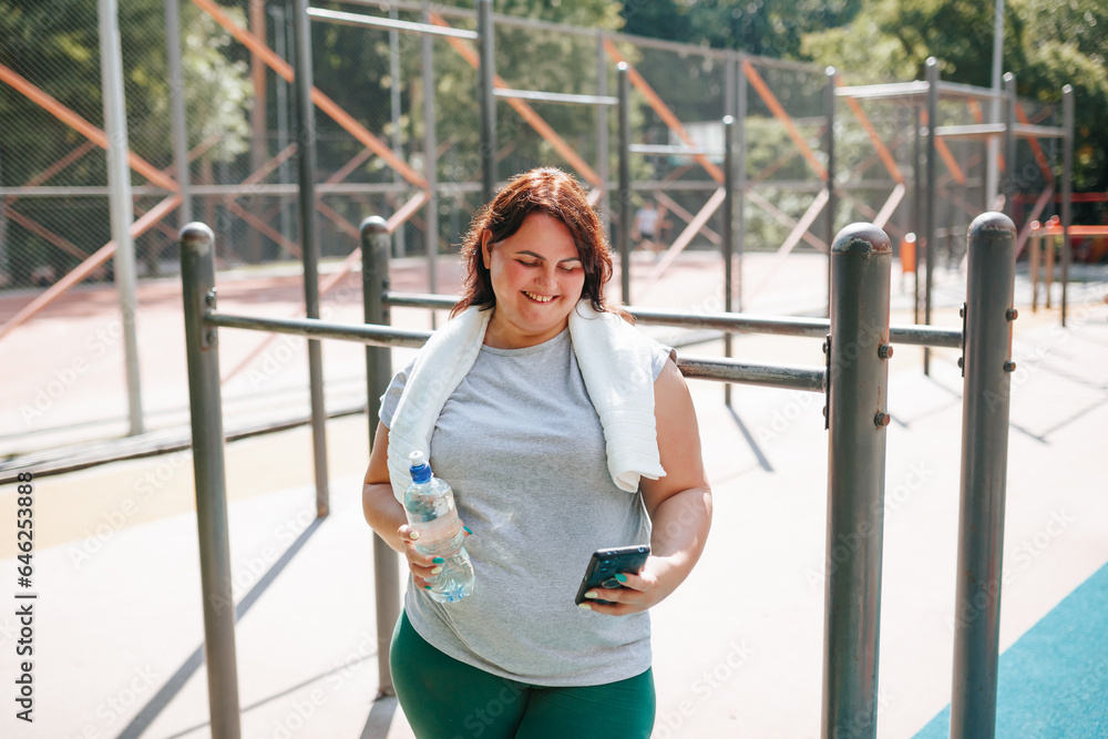 A female with plus size, after outdoor training, finds joy with her smartphone. Her smile radiates satisfaction, with a water bottle by her side.