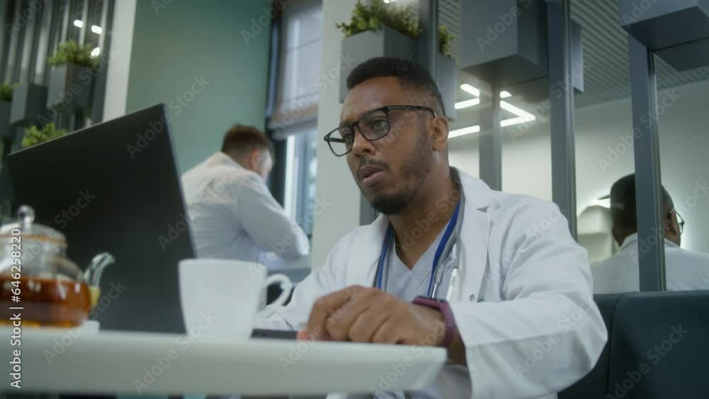 African American doctor uses laptop in modern clinic cafe during break. Medic drinks tea or coffee, watches medical tests results of patient or surfs the Internet. Medical staff in hospital cafeteria. African American doctor uses laptop in modern clinic cafe during break. Medic drinks tea or coffee, watches medical tests results of patient or surfs the Internet. Medical staff in hospital cafeteria.