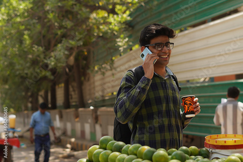 Young Indian middle class young man holding glass in hand and talking on smartphone