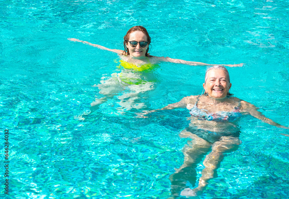 Couple of two mature women friends having fun floating in outdoor swimming pool under the sun. Vacation, healthy lifestyle and relax concept