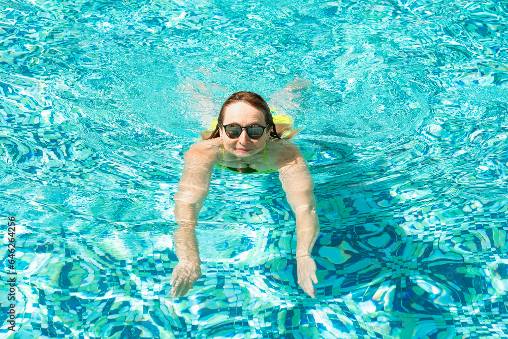 Active mature caucasian woman swimming in outdoor swimming pool under the sunlight wearing sunglasses. Vacation and healthy lifestyle concept