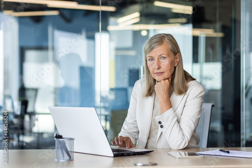 Portrait of serious mature thinking businesswoman, gray haired female boss looking at camera, at workplace inside office, financier with laptop, confident accountant investor.
