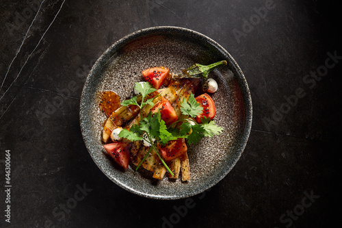 Horizontal top view of roasted eggplant with tomatoes, cilantro, and cheese on a stone plate, set against a minimalist black marble background, embodying pan-Asian flavors