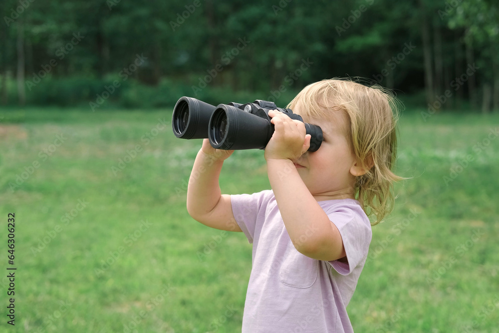 Little girl using binoculars in a forest. 2 years kid looking ahead. Smiling baby with spyglass. Adventure, Imagination, travel concept. Freedom, vacation. Happy child playing outdoor in summer field