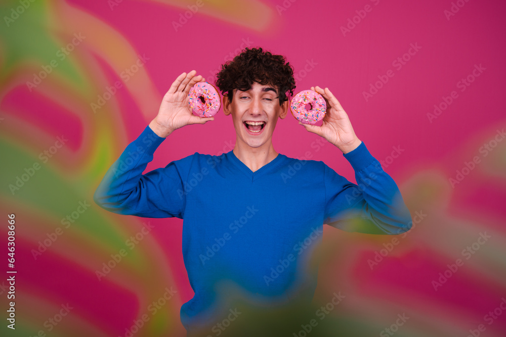 Young attractive curly man eating pink donuts and posing on a pink background. Young attractive curly man eating pink donuts and posing on a pink background.