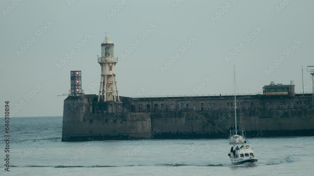 Small boat enters the harbour of Dover England with the seawall and lighthouse visible. A yacht is sailing behind Small boat enters the harbour of Dover England with the seawall and lighthouse visible. A yacht is sailing behind