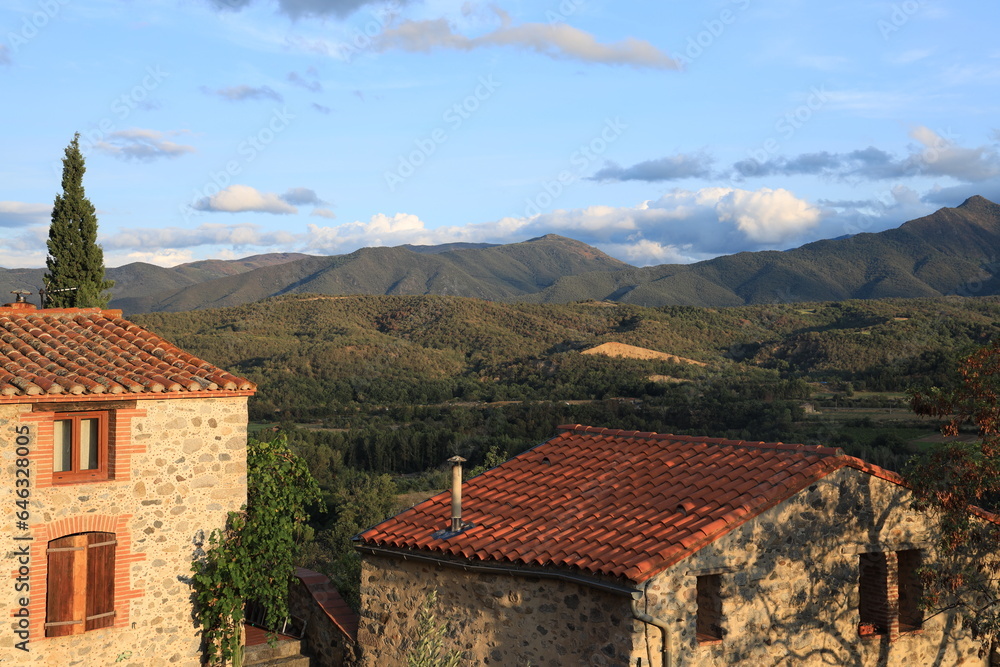 View of traditional stone houses with terra cotta roof tiles in evening sunlight with sprawling hill countryside visible in background. Eus, southern France