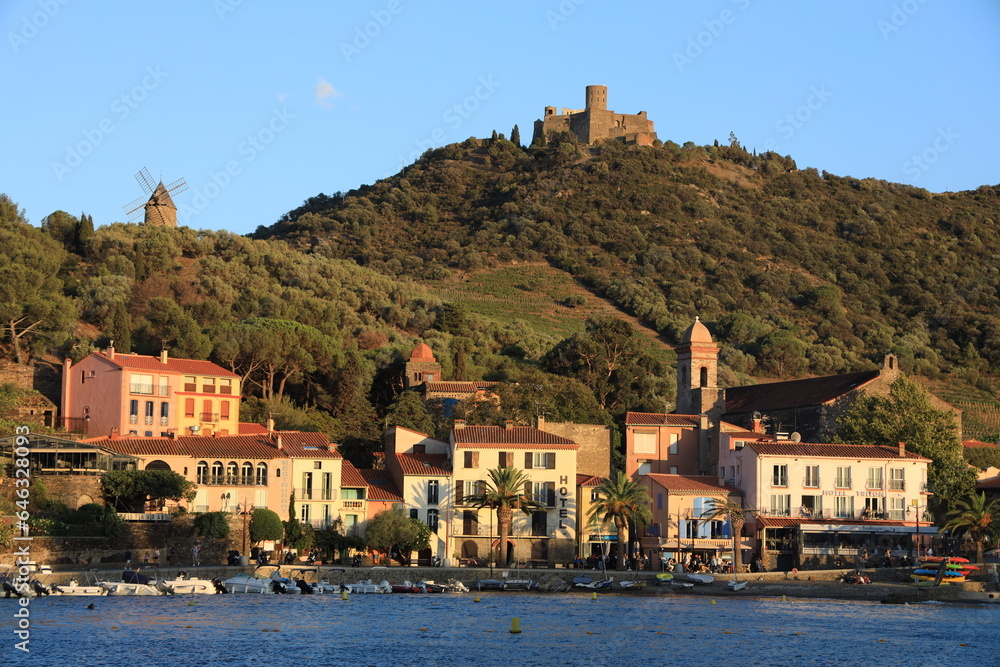 Collioure, France - August 2023: a view of Collioure from across bay featuring brightly coloured buildings at foot of hill with windmill and Fort Saint Elme visible at top of hill
