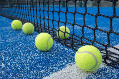 Four yellow balls on floor in front and one behind of paddle net in blue court outdoors. Padel tennis © Vic