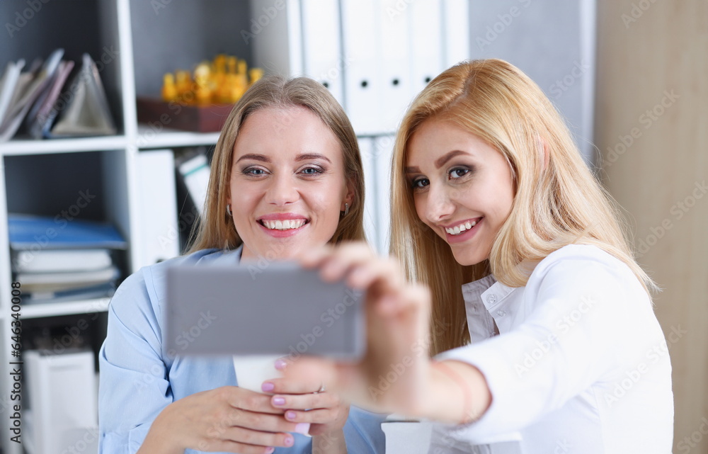 Two businesswomen in the office smile and do selfie on a smartphone during a break to have coffee Two businesswomen in the office smile and do selfie on a smartphone during a break to have coffee