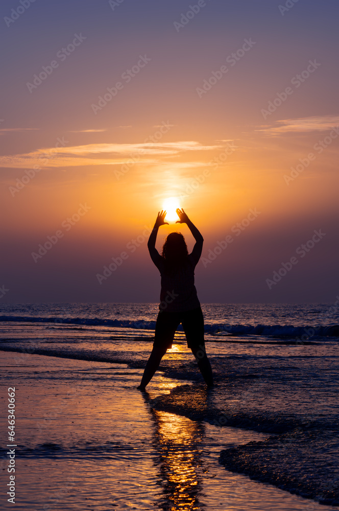 Silhouette of girl reaching for the sun on the beach of a frisian island on the North Sea coast in Germany. Colorful sunset sky reflected by waves and surf. Summer greetings from holiday destination.
