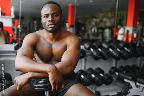 Hot african american young man bodybuilder lifting barbell at gym, working on his arms, looking at copy space. Black muscular shirtless guy having biceps workout session. Healthy lifestyle concept.