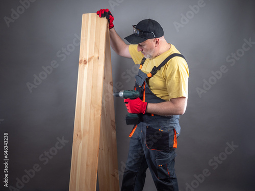 Furniture assembler with boards. Man uses electric screwdriver. Worker in gray suit. Builder with screwdriver. Construction company worker. Man carpenter on black. Master with parquet board