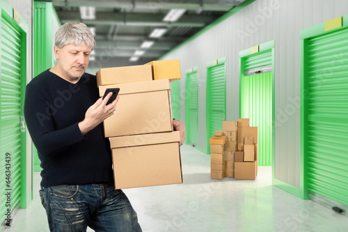 Storage room tenant. Man in warehouse building. Tenant with boxes and phone. Gray-haired man rents warehouse. Guy in corridors of storage company. Male with phone at entrance to storage room © Grispb