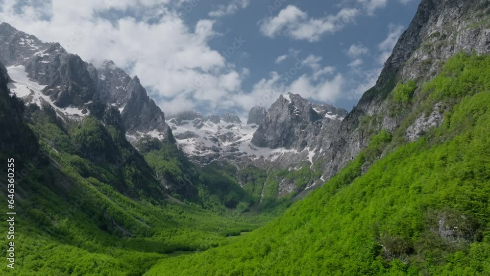 Establishing aerial view huge mountains in early summer with snowy peaks, blue sky with clouds and green valley with a forest. Establishing aerial view huge mountains in early summer with snowy peaks, blue sky with clouds and green valley with a forest.
