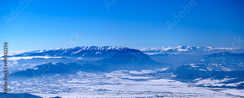Incredible winter landscape of alpine valley under bright sunny light in frosty morning, beautiful alpine panoramic view of snow capped Piatra Craiului and Fagaras mountains in background, Romania