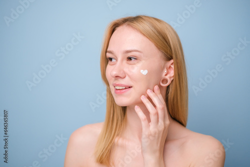 A beautiful girl applies moisturizer to her facial skin. Young woman on blue background smiling and taking care of her skin.