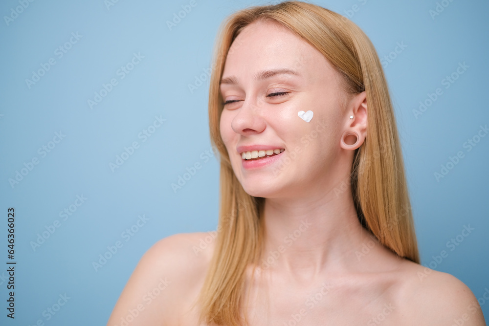 A beautiful girl applies moisturizer to her facial skin. Young woman on blue background smiling and taking care of her skin.