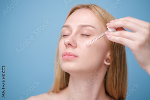 Beautiful girl applying serum on her face standing on blue background. Young woman holding a dropper with skin care product for healthy and glowing skin color.