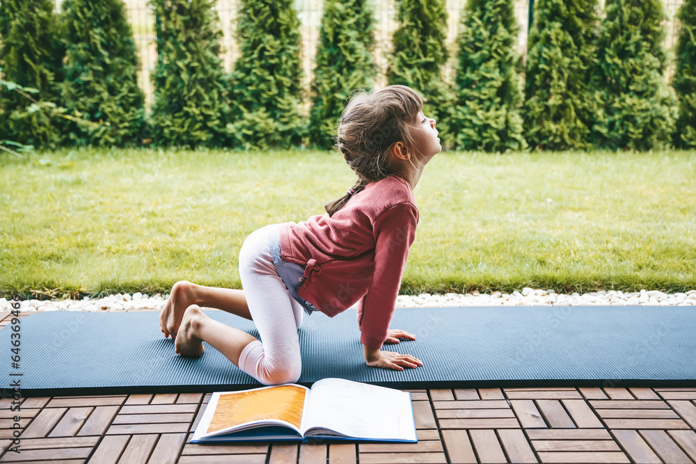 Cute little girl 5-6 years old standing in Marjariasana or Cat pose on the roll mat practicing yoga outdoors