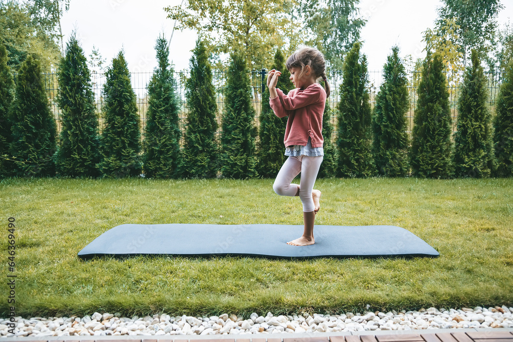 Naklejka premium Cute little girl 5-6 years old standing in the Eagle position or Garudasana while practicing yoga outdoors