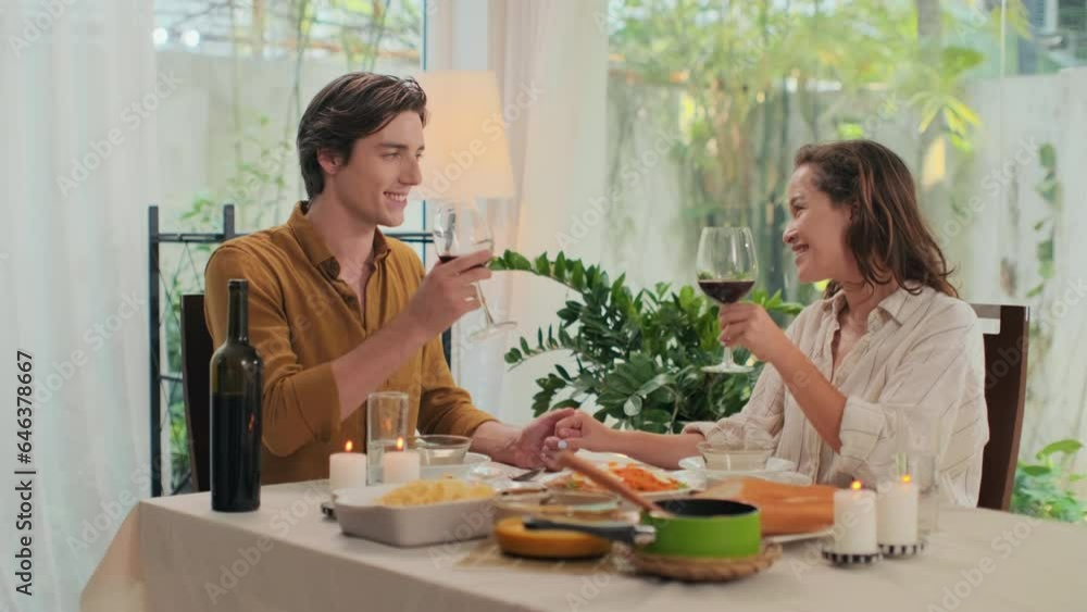 Medium shot of young intercultural couple clinking glasses with red wine when celebrating anniversary at romantic dinner at home Medium shot of young intercultural couple clinking glasses with red wine when celebrating anniversary at romantic dinner at home