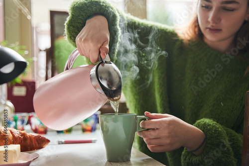 Calm teen girl pouring hot tea while sitting at kitchen during breakfast © Yakobchuk Olena