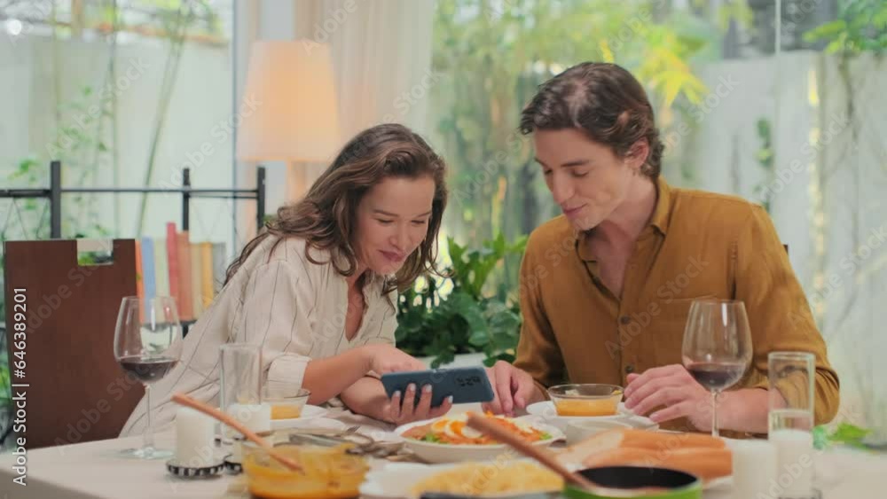 Medium shot of young cheerful couple eating puree for romantic dinner and watching funny videos on smartphone while sitting at festive table at home Medium shot of young cheerful couple eating puree for romantic dinner and watching funny videos on smartphone while sitting at festive table at home