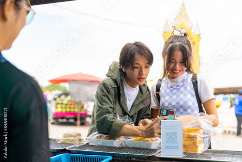 Group of Happy Asian people tourist buying and eating street food with using mobile app on smartphone scan QR code making online payment during travel local town in Thailand on summer holiday vacation photo