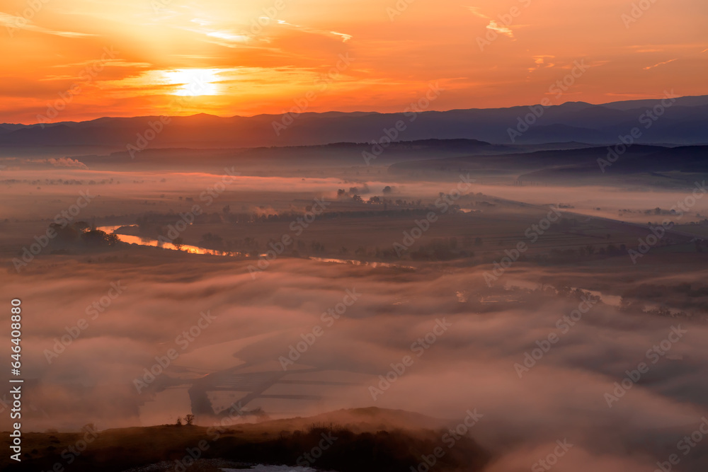 Fototapeta premium Aerial view of the sunrise over the valley in beautiful early morning mist in the highlands. Low clouds and fog cover the sleeping meadow. Hills valley mists landscape. Serene moment in rural area