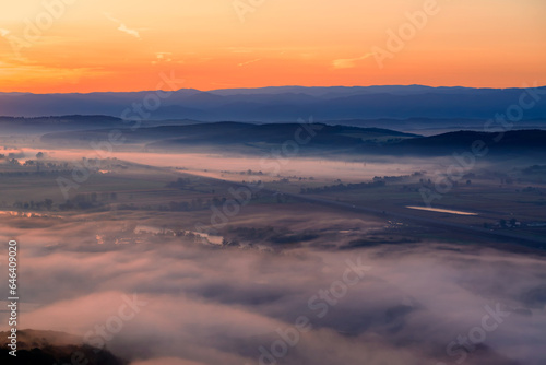 Aerial view of the valley in early morning mist  beautiful in the highlands. Low clouds and fog cover the sleeping meadow. Alpine mountain valley mists landscape at dawn. Serene moment in rural area