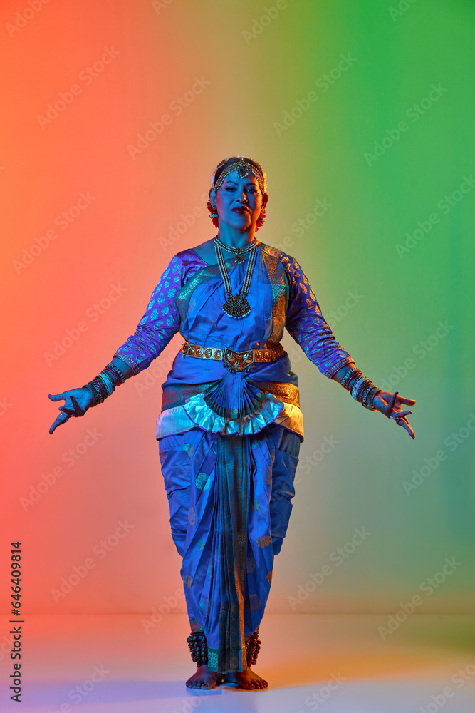 Fototapeta premium Beautiful indian woman dancing traditional dance in elegant blue dress against gradient studio background in neon light. Concept of beauty, fashion, India, traditions, choreography, art. Ad
