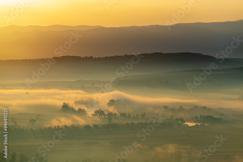 Fototapeta Naklejka Na Ścianę i Meble -  Aerial view of the valley in early morning mist, beautiful in the highlands. Low clouds and fog cover the sleeping meadow. Alpine mountain valley mists landscape at dawn. Serene moment in rural area