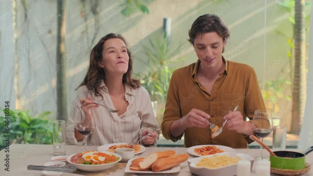 Medium shot of intercultural couple eating pasta bolognese while sitting at dinner table with glasses of red wine Medium shot of intercultural couple eating pasta bolognese while sitting at dinner table with glasses of red wine