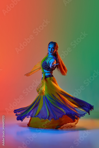 Following asian tradition. Woman in indian dress, makeup and accessories dancing against gradient studio background in neon light. Concept of beauty, fashion, India, traditions, choreography, art. Ad