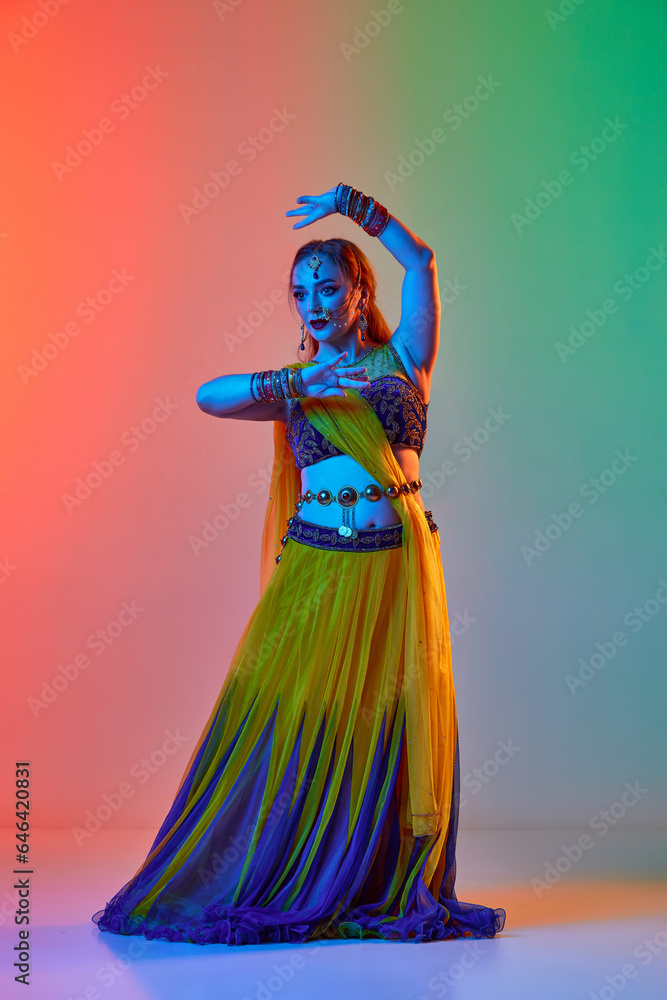 Fototapeta premium Odissi dance. Beautiful young woman in traditional dress dancing against gradient studio background in neon light. Concept of beauty, fashion, India, traditions, lifestyle, choreography, art. Ad