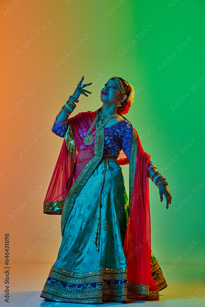 Elegant, mature, beautiful indian woman in traditional clothes, dress posing against gradient studio background in neon light. Concept of beauty, fashion, India, traditions, choreography, art. Ad