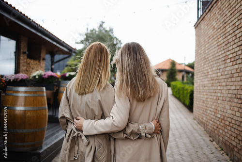 Two friends, a woman with long hair blonde. Concept: friendship, twins, autumn, weekend vacation in the fresh air.. Rear view, sister girls, in stylish beige trench coats, stand hugging each other.