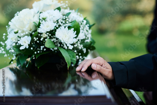 Focus on hand of mourning mature woman in black attire on lid of closed wooden coffin with bunch of fresh white chrysanthemums on top photo