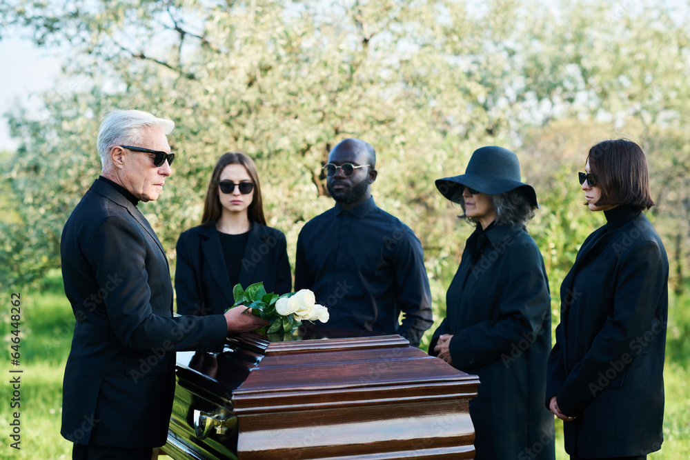 Mature man in black suit and sunglasses putting bunch of white roses on