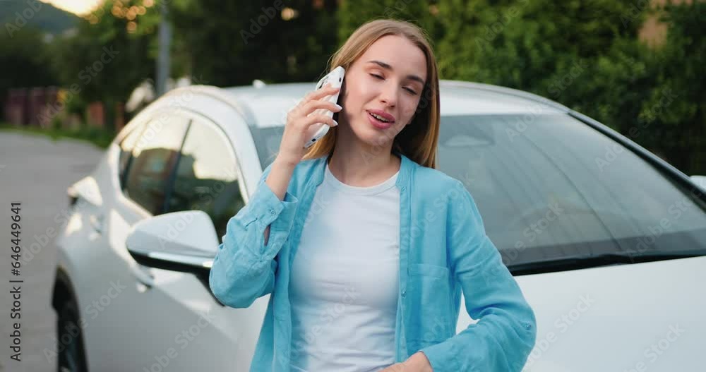 Beautiful young woman talking on cell phone answering friends call when leans on her white car which is parked by area house. Outdoors Beautiful young woman talking on cell phone answering friends call when leans on her white car which is parked by area house. Outdoors