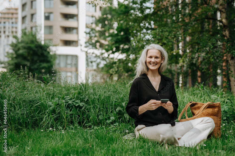 Portrait of beautiful woman with gray hair sitting on the grass in city park, using her smartphone. Portrait of beautiful woman with gray hair sitting on the grass in city park, using her smartphone.