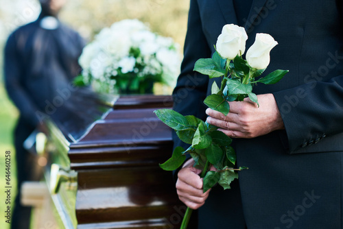 Close-up of matue man in black suit holding two fresh white roses while standing in front of camera against coffin with closed lid