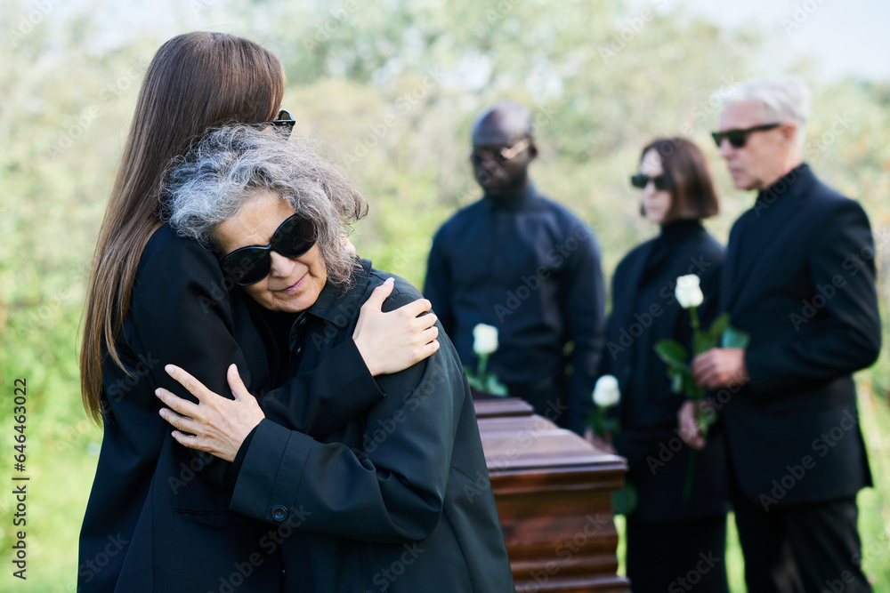 Young grieving woman giving hug to her mourning mother in sunglasses crying on her shoulder while standing against coffin and group of people