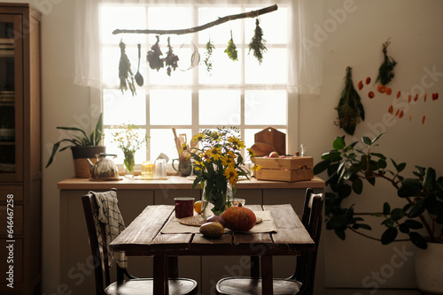 Two wooden chairs standing by table with bunch of yellow garden flowers in vase  ripe pumpkin and eggplants and mug against kitchen counter